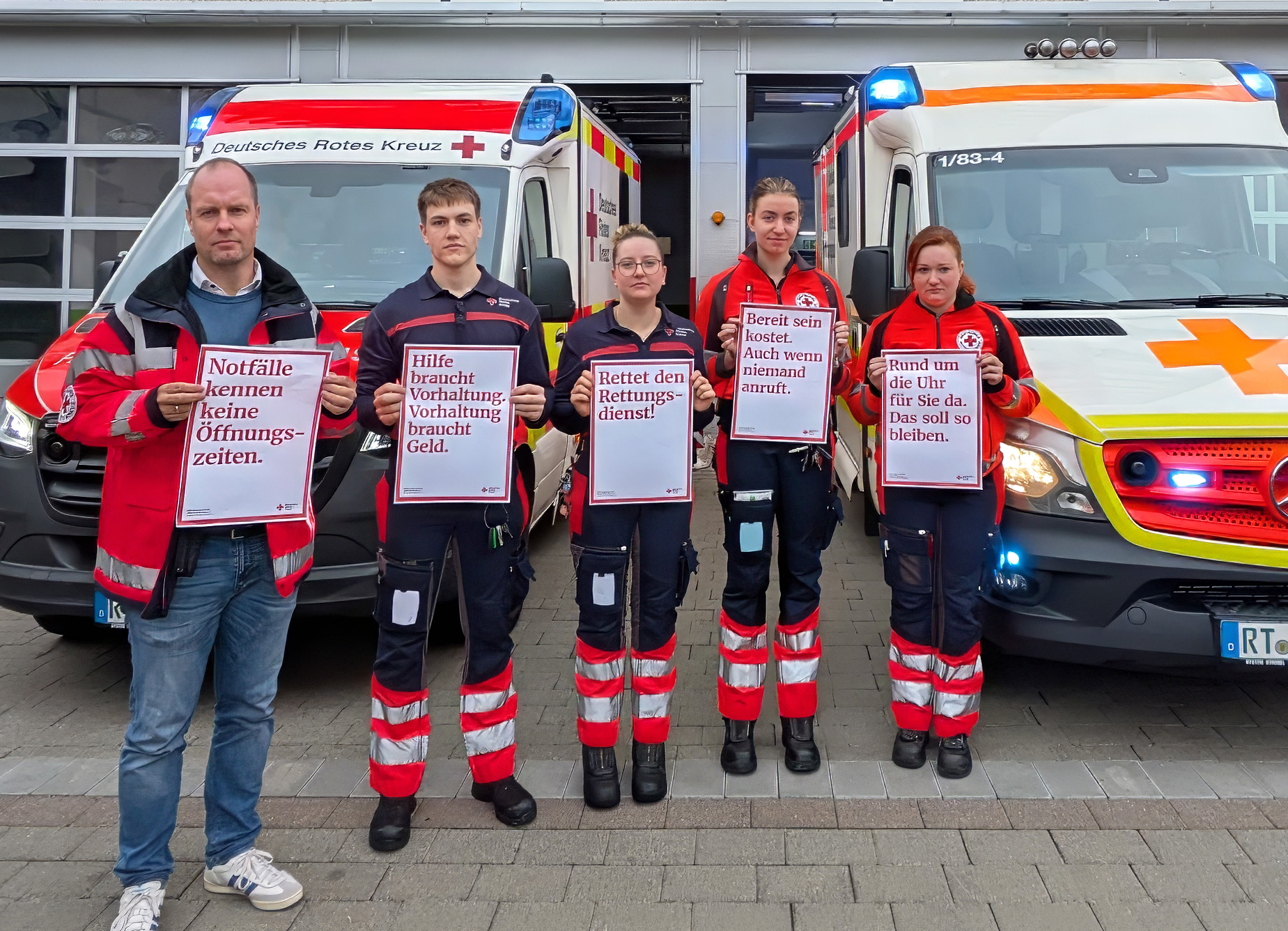 Bild: DRK-Kreisgeschäftsführer mit Mitarbeitenden aus dem Rettungsdienst stehen mit Plakaten vor 2 Einsatzfahrzeuge